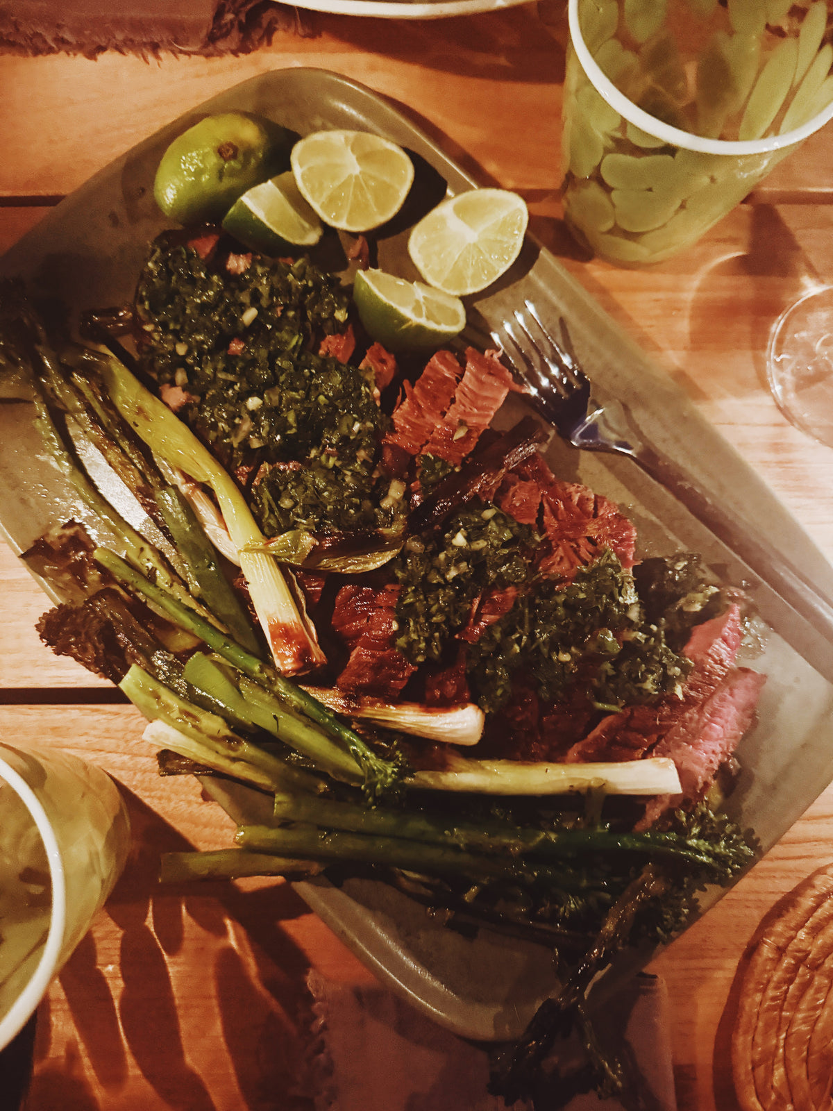 A platter of sliced steak under chimichurri, with charred scallions and limes