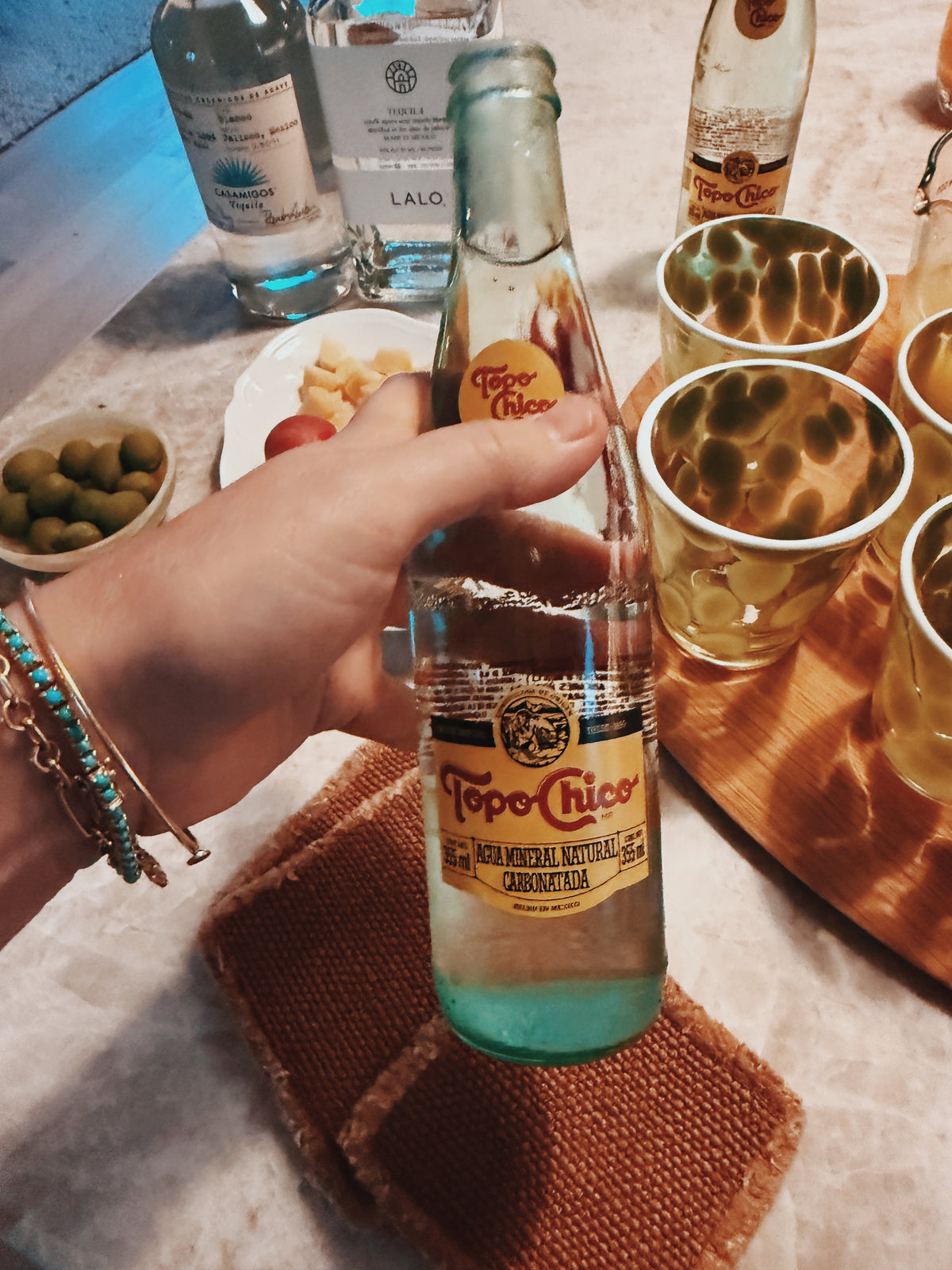 A hand holding a bottle of Topo Chico over a tray of glasses and tequila
