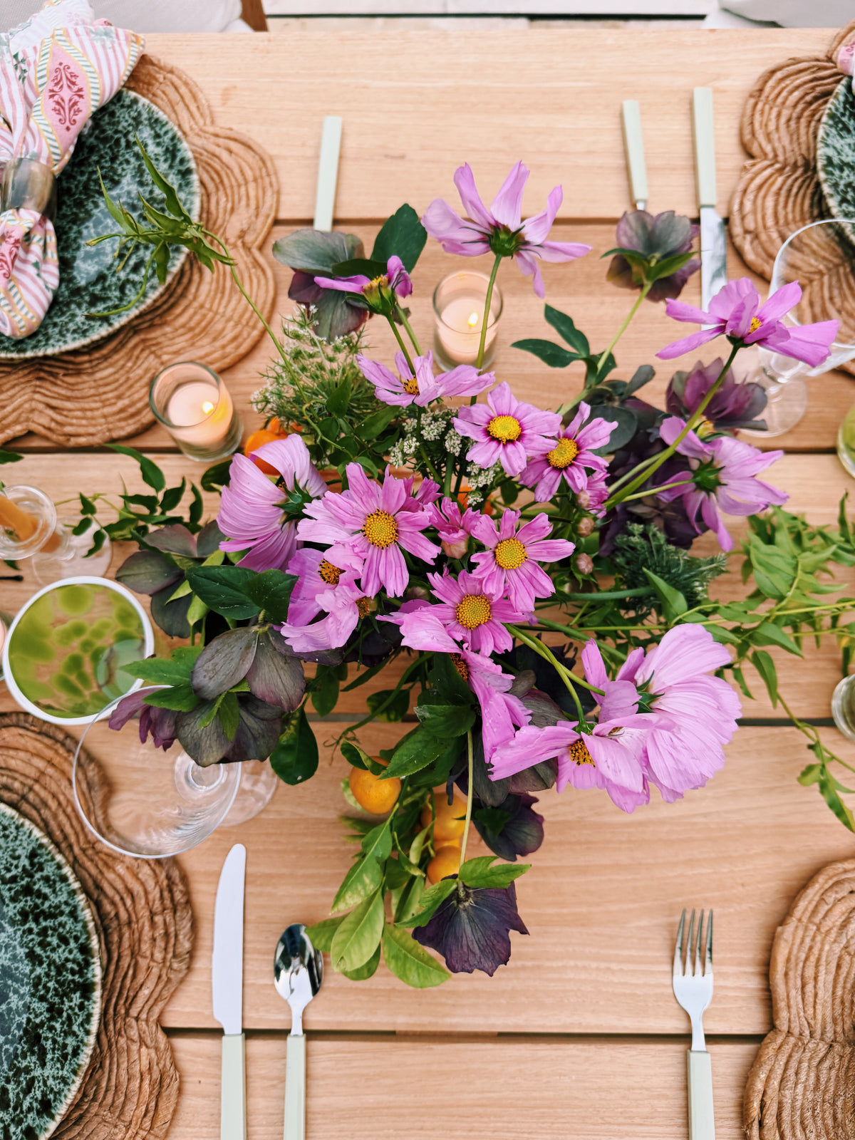 Pink cosmos and greenery in a low arrangement on a wooden table, with woven chargers and green plates