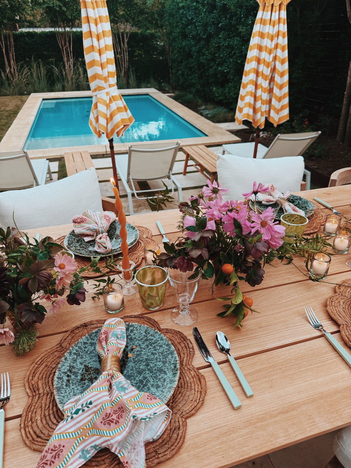 A long table laid outside by the pool, with woven chargers, pink flowers and striped umbrellas