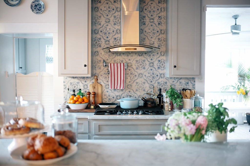 A kitchen with blue and white tiles behind the stove, a bowl of oranges, a cloche of pastries and a pot of pink flowers on the marble counter