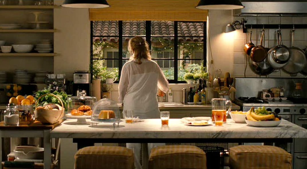 A long, sunny film-set kitchen with an island laid with breakfast, copper pans hanging at the back