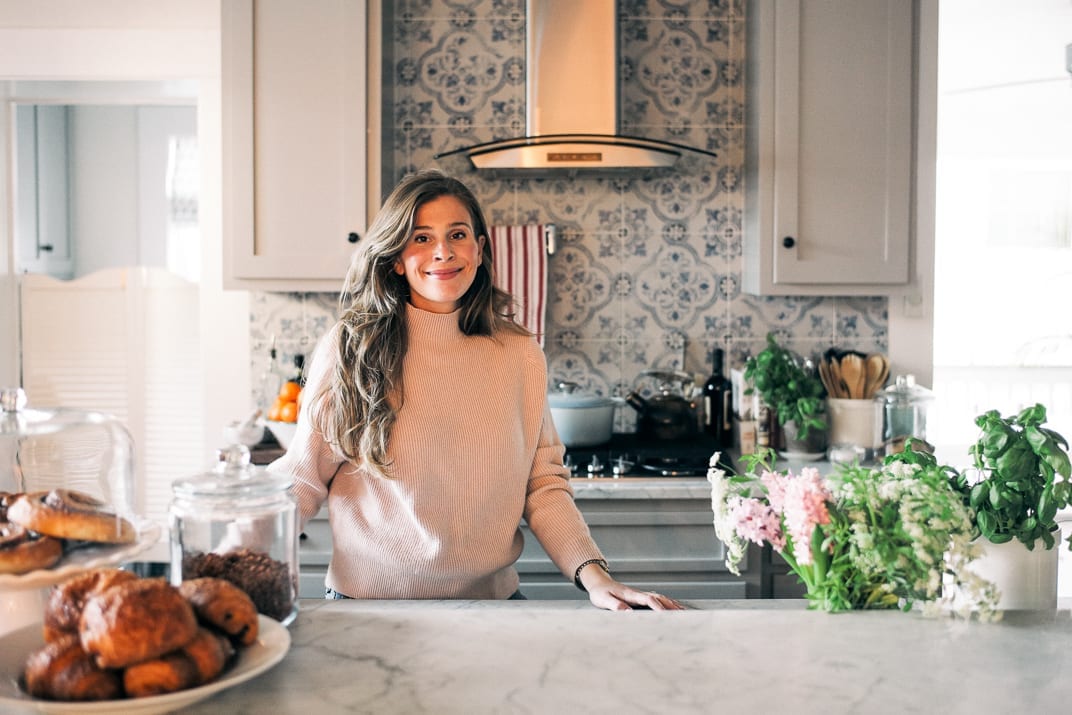 Lucy at her marble kitchen counter with blue and white tiles behind, pastries under a cloche and pink flowers