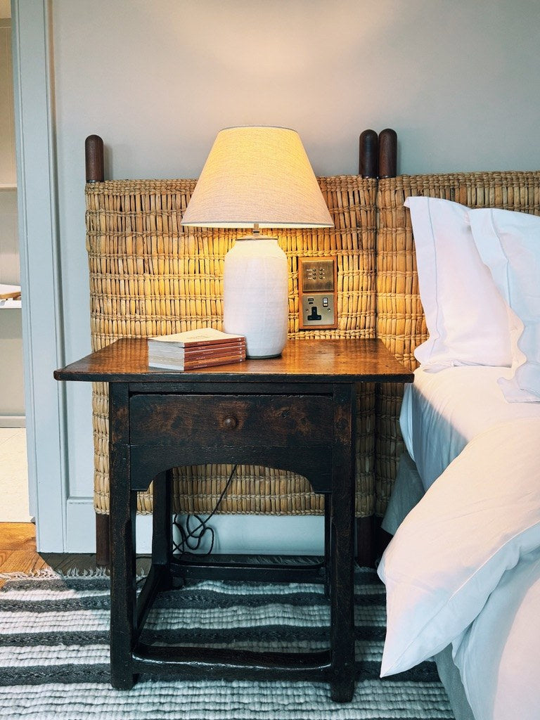 A woven headboard and a lamp on a dark bedside table