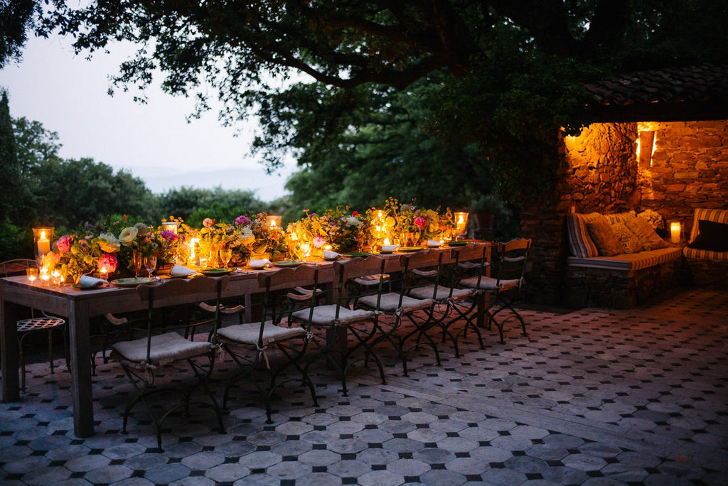 A long table on the terrace at dusk, flowers and candles from end to end, the stone sitting nook glowing beside it