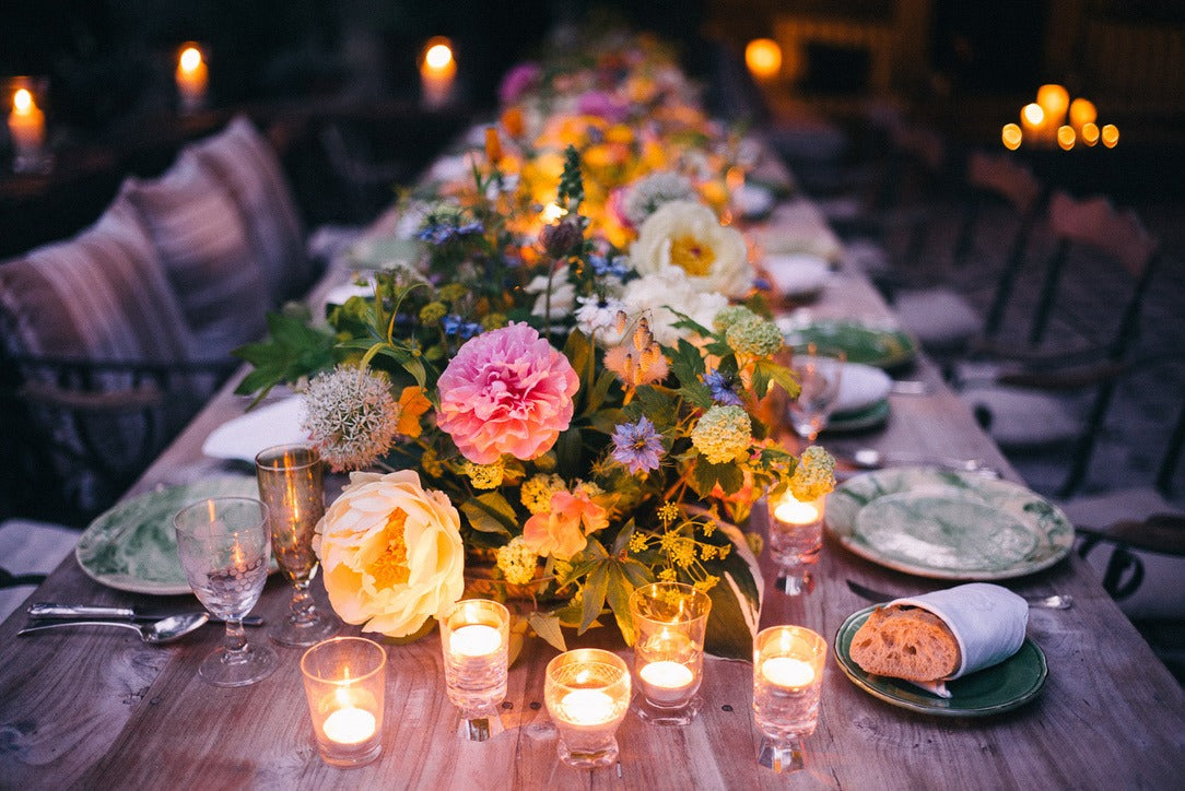 Lucy lighting the table at dusk, tapers and votives among low red flowers, the house glowing behind