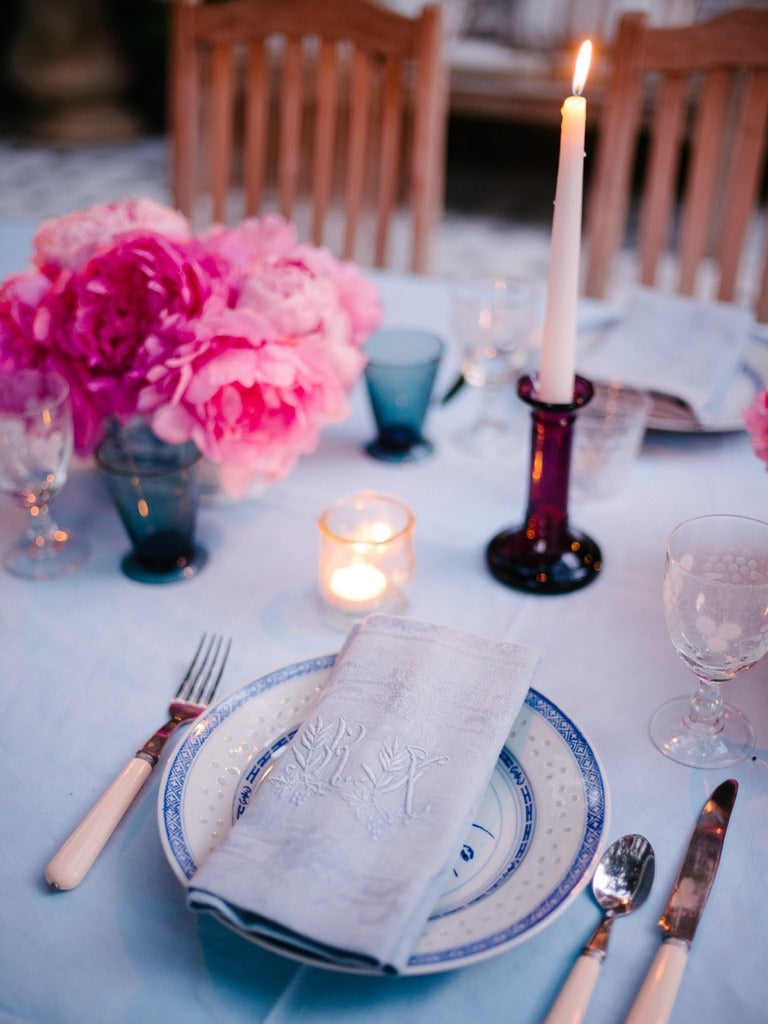 A table laid in white and blue under the vines, white roses and peonies down the middle, cushions on the bench behind