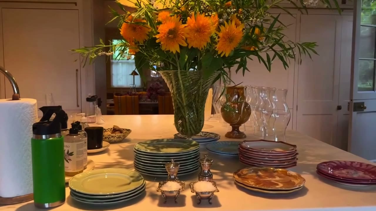 Stacks of collected plates beside a big jug of sunflowers on a kitchen counter