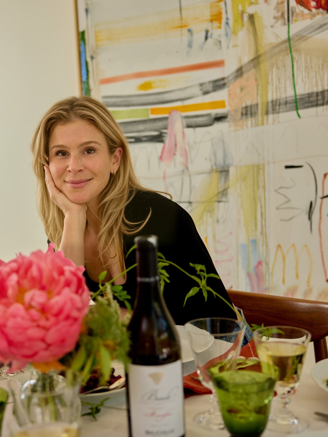 Lucy at a candlelit table, chin in hand, peonies and a bottle of wine in front of her